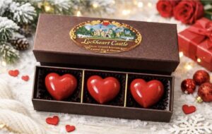 ロックハート城のロゴが入った高級感のあるブラウンのボックスに並ぶ、宝石のように真っ赤な3つのハート型チョコレート。背景には雪やクリスマスオーナメントがあしらわれたロマンチックな演出。