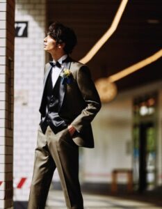 クラシックなタイルの壁の前で、光を浴びて佇む新郎。光沢のあるカーキ系のタキシードに黒のベスト、ボウタイ、イエローのブートニアを合わせた、ロックハート城の雰囲気にふさわしい洗練された着こなし。