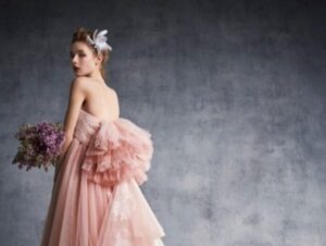 背中の大きなフリルが特徴的な、くすみピンクのカラードレスを纏った新婦のバックスタイル。ロックハート城の重厚な石壁に映える、フェザーのヘッドドレスとパープルのブーケを合わせたスタイリッシュなコーディネート。