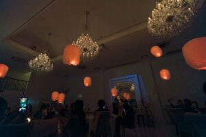 群馬県「ロックハート城」の披露宴会場で、ゲストと共に楽しむランタン飛ばしの演出。高い天井の豪華なクリスタルシャンデリアの間を、温かいオレンジ色の光を放つスカイランタンがゆっくりと舞い上がる幻想的な夜の結婚式のワンシーン。