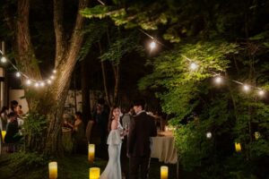 深い緑に囲まれた夜のガーデンで、ゲストとグラスを片手に談笑する新郎新婦。頭上に輝く電飾と足元のキャンドルが、**「キンセンカ(カレンデュラ)」**が閉じ込めた太陽の光のように温かく周囲を照らしている。ロックハート城の森の中で、飾らない自然体な幸せを分かち合う、陽だまりのような結婚式のワンシーン。