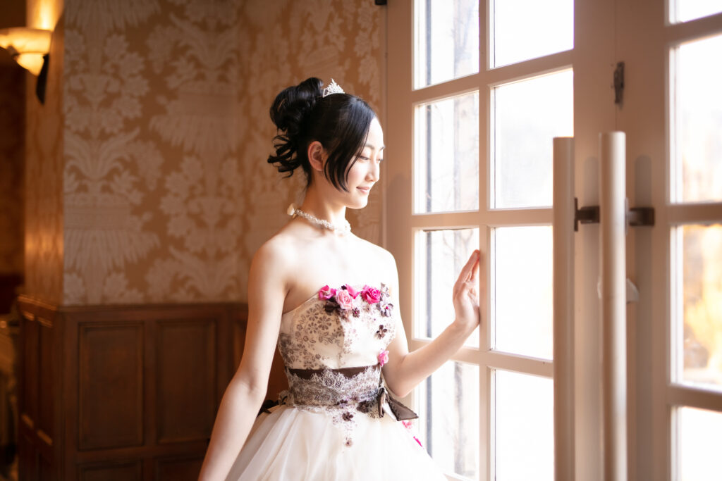 「アンティーク調の窓辺で、花飾りのついた白いドレスを纏い、柔らかな光に包まれる女性（プリンセス体験）」