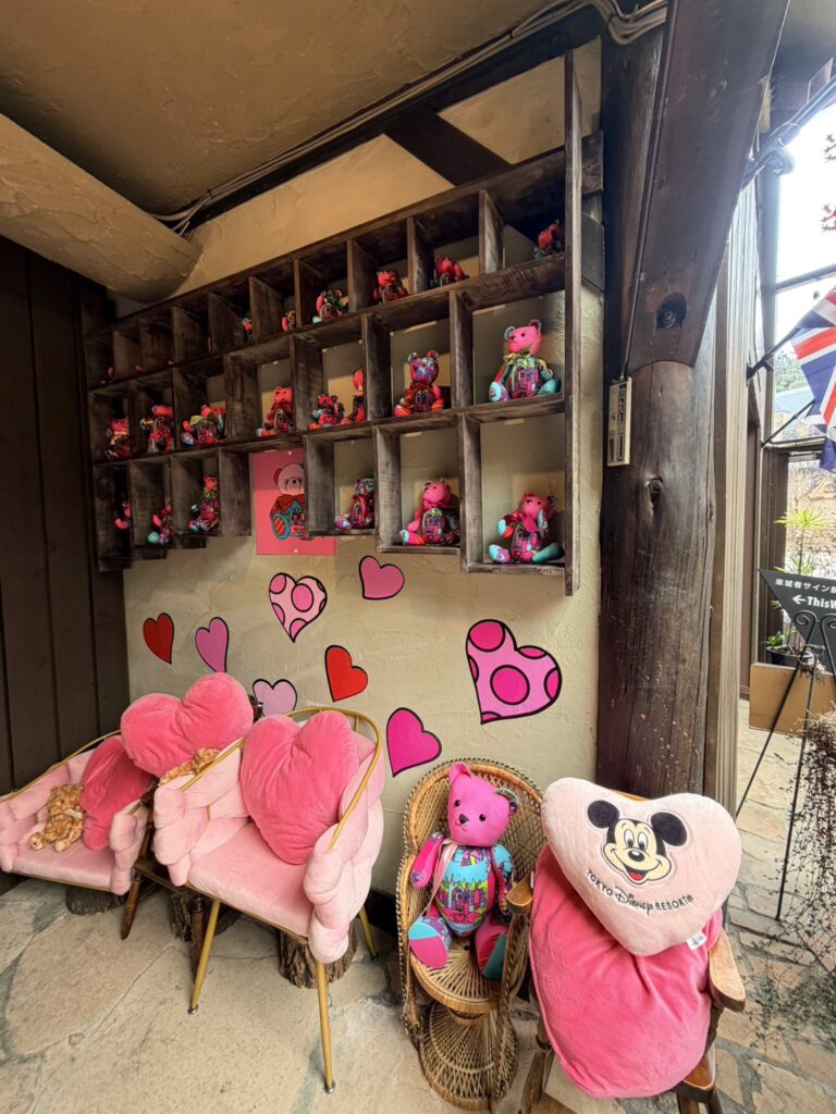 木製の棚に並ぶ色鮮やかなピンクのテディベアと、壁に描かれたハートのイラスト、ピンクのハート型クッションが置かれた華やかなフォトスポット。