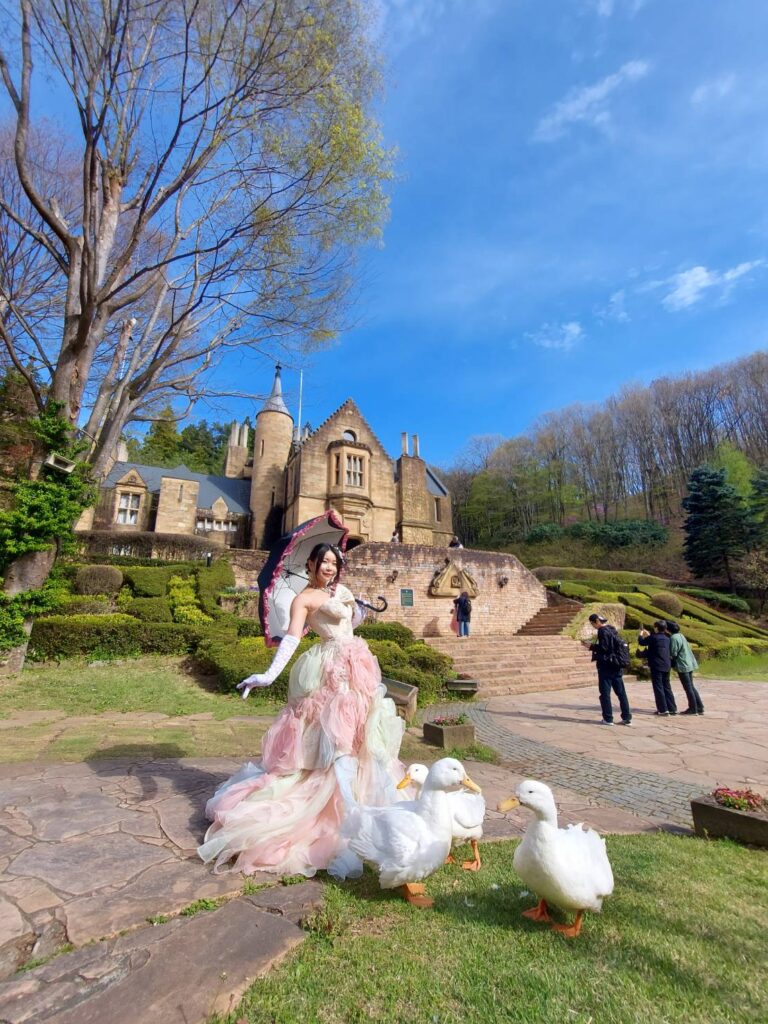 ドレスを着た女性が石造りの洋館を背景に、可愛らしいアヒルたちとポーズをとっている様子。