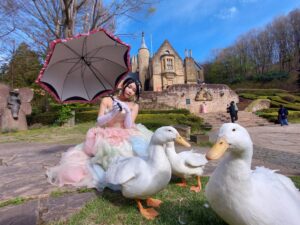 ドレスを着た女性が石造りの洋館を背景に、可愛らしいアヒルたちとポーズをとっている様子。