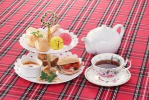 赤いタータンチェックのテーブルクロスの上に置かれた、2段式のティースタンド（マカロン、シュークリーム、サンドイッチ、スコーンなど）と白いティーポット、花柄のカップに注がれた紅茶。群馬県高山村、ロックハート城