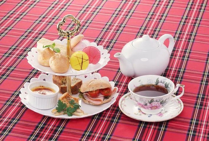 赤いタータンチェックのテーブルクロスの上に置かれた、2段式のティースタンド(マカロン、シュークリーム、サンドイッチ、スコーンなど)と白いティーポット、花柄のカップに注がれた紅茶。