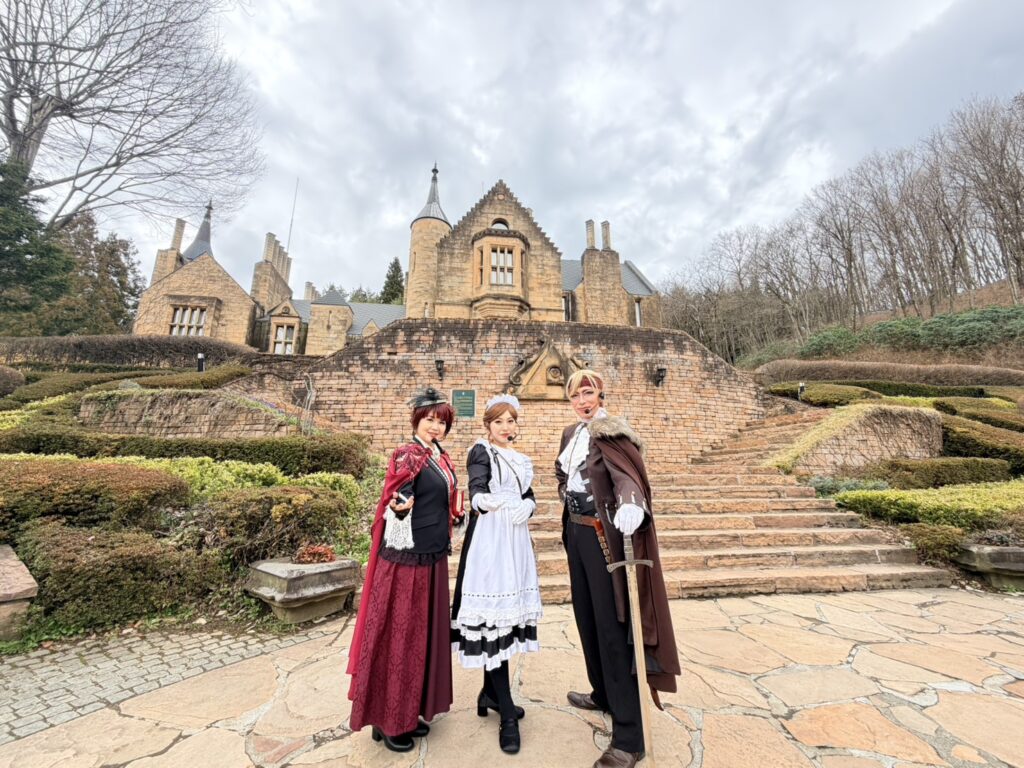 ロックハート城の階段前で、華やかな衣装に身を包んだダグラスバトラーズの3名がお客様を歓迎している様子。左には深紅のドレスの執事、中央には可憐なメイド、右には剣を携えたマント姿の騎士が並び、非日常的な世界観を演出している。