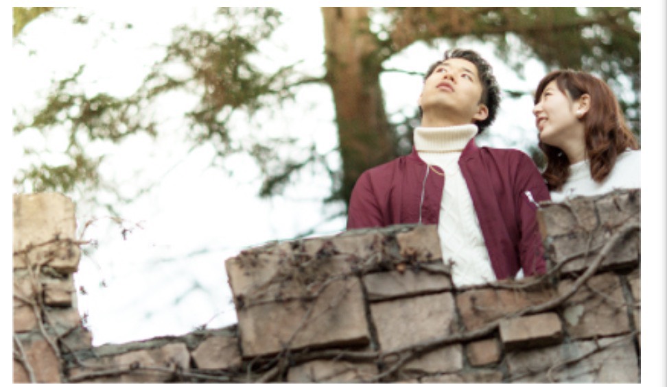 緑豊かな庭園の石壁から、空を見上げる男性と、隣で優しく微笑む女性のナチュラルなペアフォト。