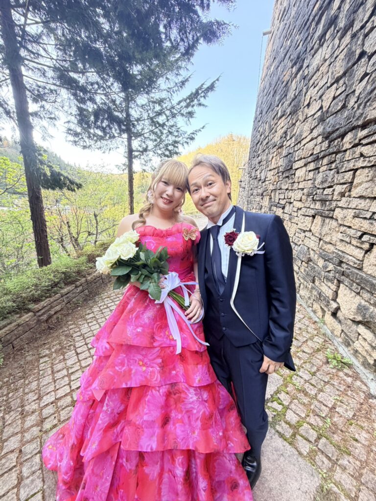 豊かな緑と石壁のあるガーデンで、鮮やかなピンクのウェディングドレスを着た花嫁と、ネイビーのタキシードを着た花婿が、白いバラのブーケを持って仲良く並んで立っている写真。