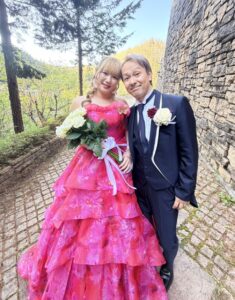 豊かな緑と石壁のあるガーデンで、鮮やかなピンクのウェディングドレスを着た花嫁と、ネイビーのタキシードを着た花婿が、白いバラのブーケを持って仲良く並んで立っている写真。