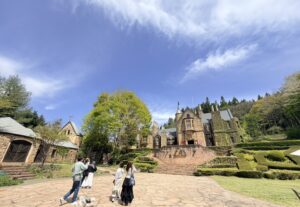 青空の下、石畳の広場の奥に建つ、ツタの絡まる中世ヨーロッパ風の石造りの大きな洋館。手前には庭園と階段、そして数人の観光客が写っている。