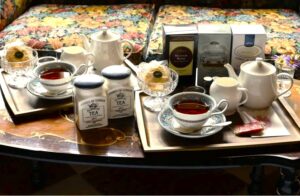 上品なティーカップ、白いティーポット、スコーンが並んだ2名分のティーセット。背景には「ASHBYS OF LONDON（アシュビィズ・オブ・ロンドン）」の紅茶缶やパッケージが並び、英国式の本格的なティータイムを演出している様子。