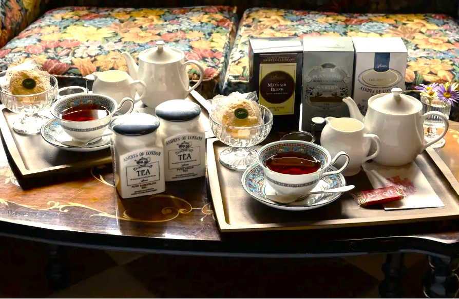 アンティーク調のテーブルに、上品なティーカップ、白いティーポット、スコーンが並んだ2名分のティーセット。背景には「ASHBYS OF LONDON（アシュビィズ・オブ・ロンドン）」の紅茶缶やパッケージが並び、英国式の本格的なティータイムを演出している様子。