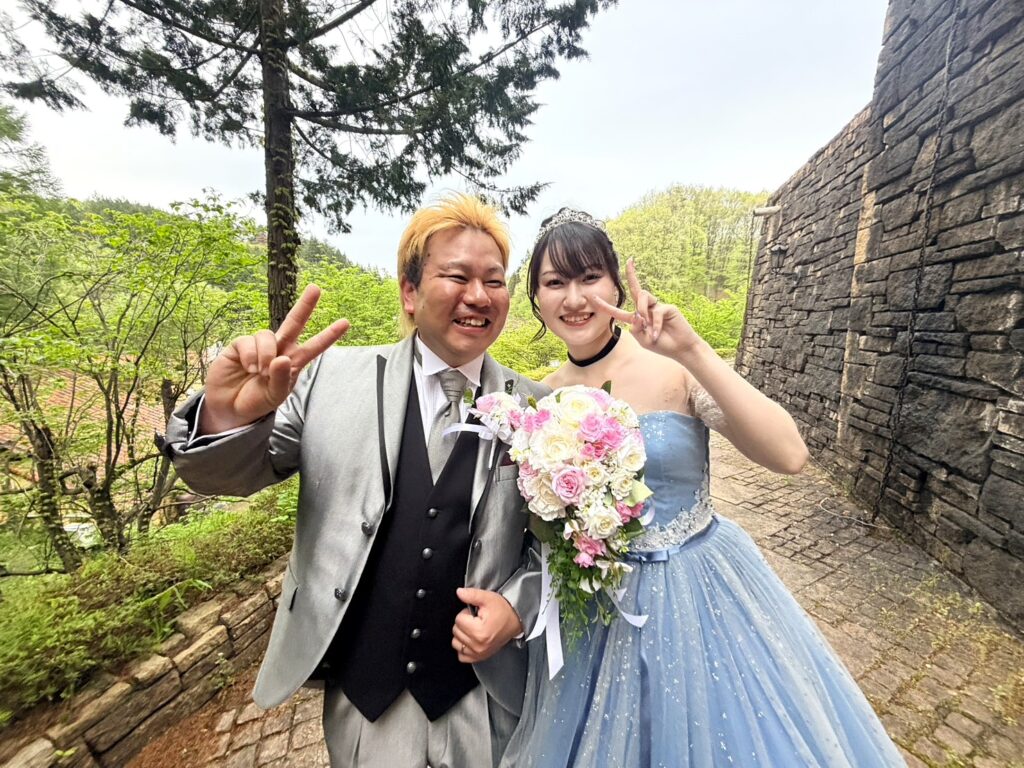 ロックハート城の豊かな緑を背景に、笑顔でピースサインをするタキシード姿の新郎と水色のカラードレスを着た新婦。
