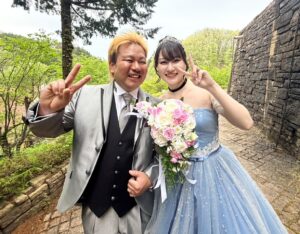 ロックハート城の豊かな緑を背景に、笑顔でピースサインをするタキシード姿の新郎と水色のカラードレスを着た新婦。