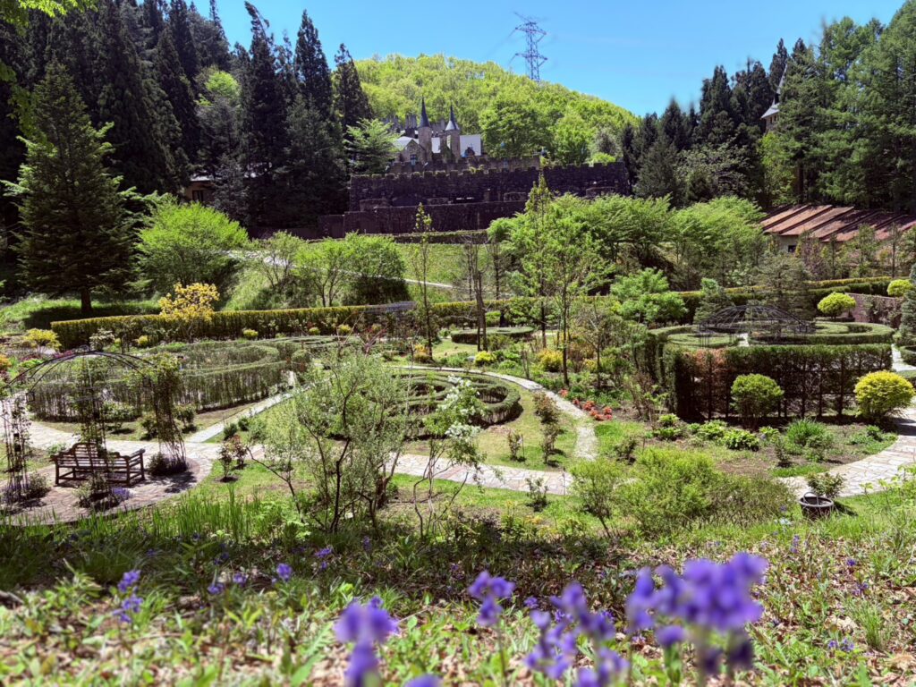 新緑の森に囲まれた古城を背景に、手前にラベンダーやハーブガーデンが広がる初夏の庭園の風景