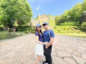 青い帽子とTシャツを合わせたカップルが、新緑に囲まれた石造りの洋館の前で仲良く並んでいる写真。足元は石畳、背景には晴天と手入れされた庭園が広がっている。