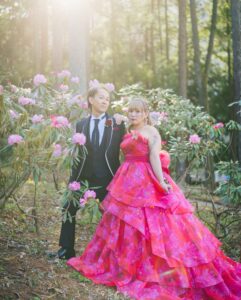 木漏れ日が差し込む森の中で、ピンクの花に囲まれて立つ、鮮やかなピンクの花柄カラードレスを着た新婦と、黒のタキシード姿の新郎のツーショット