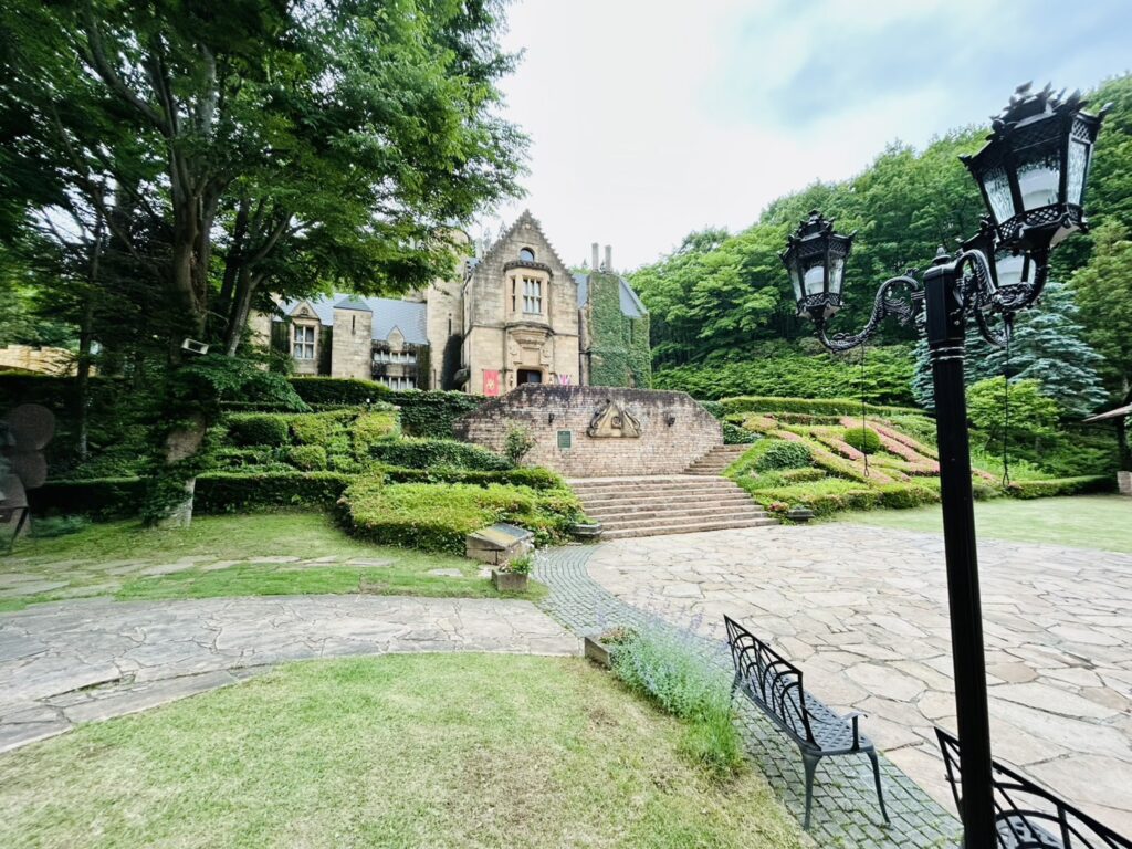 緑豊かな庭園と石畳の広場の奥に建つ、中世ヨーロッパ風の石造りの城館。右手前にはアンティーク調の黒い街灯とベンチがある。