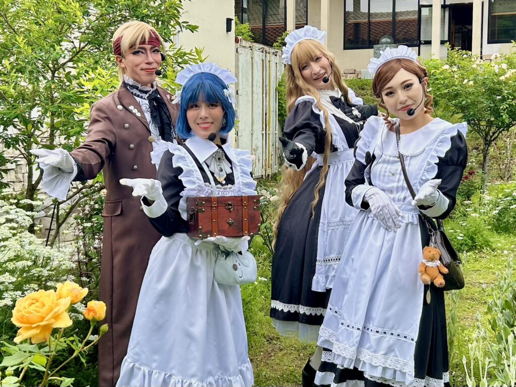 庭園で笑顔でポーズをとる、ヘッドセットマイクを装着したメイド服姿の3人と茶色の衣装を着たパフォーマー。中央の青い髪のメイドは小さな宝箱を持っています。