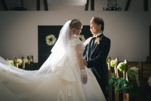 チャペルのバージンロードで、見つめ合って立つウエディングドレス姿の新婦とタキシード姿の新郎。新婦のベールとドレスに背後から光が当たっている。