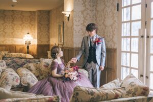 クラシックな洋室のソファでくすみパープルのカラードレスを着て微笑む新婦と、その手を取り見つめ合うシルバータキシード姿の新郎の結婚写真